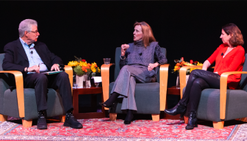 A man and two women, all wearing business casual and holding microphones, sit chatting on a stage. 