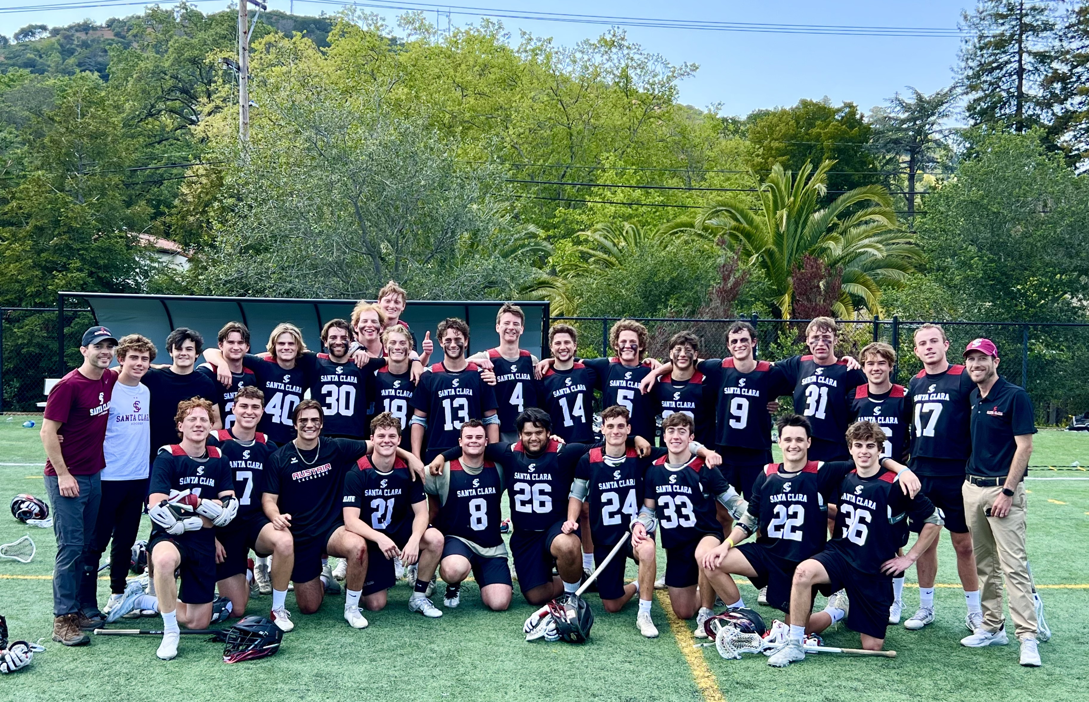 Mens lacrosse team posing on field, surrounded by trees.