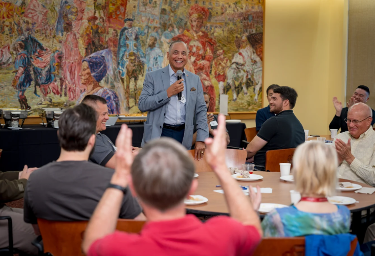 Man standing with microphone at a table while other guests clap for him