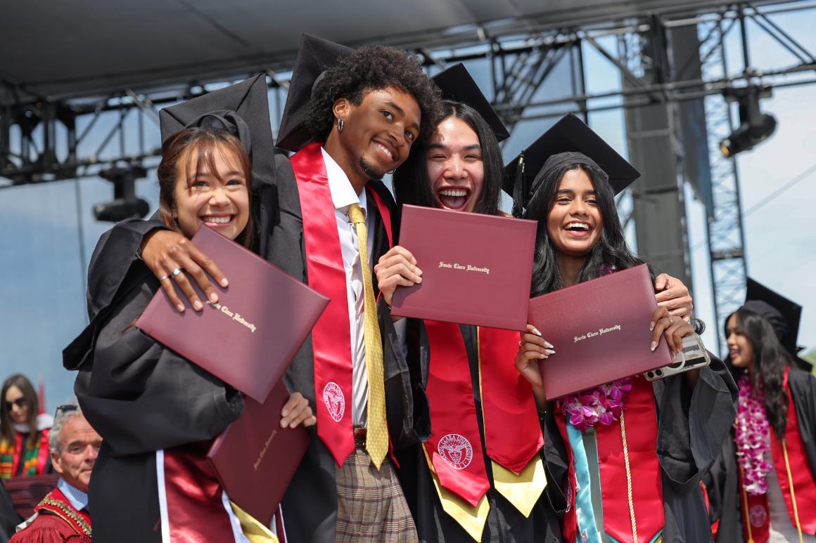 Graduates pose together on stage smiling while holding up their diplomas.