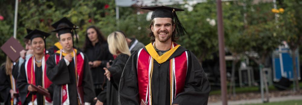 Graduates in caps and gowns walking on a red carpet stage outdoors.