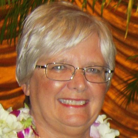 Person wearing eyeglasses and leis, smiling in front of a patterned background.