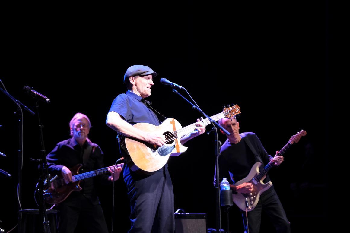 James Taylor performing on stage with a guitar and band.