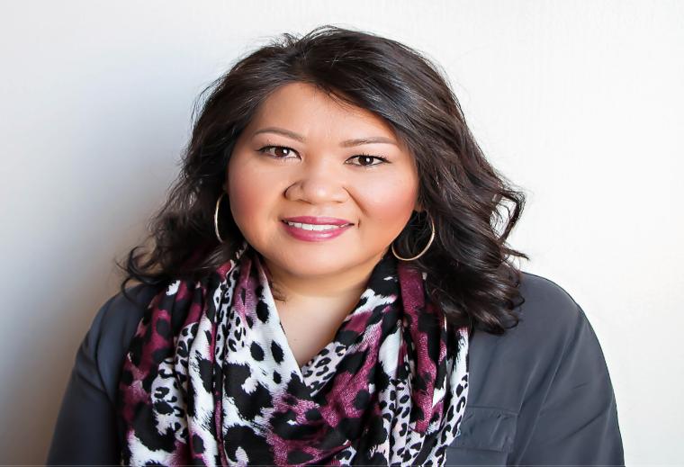 Woman wearing a patterned scarf and grey shirt smiling in front of a plain background.