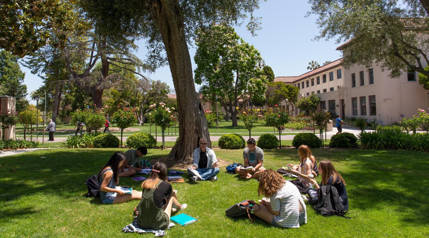 A faculty member and students sit outside in a circle discussing and taking notes