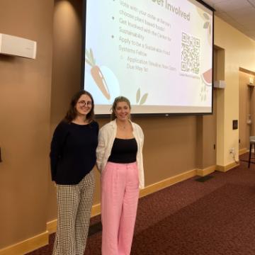 Two students stand beside a projector screen that displays their research on Food Services and Food Security at SCU