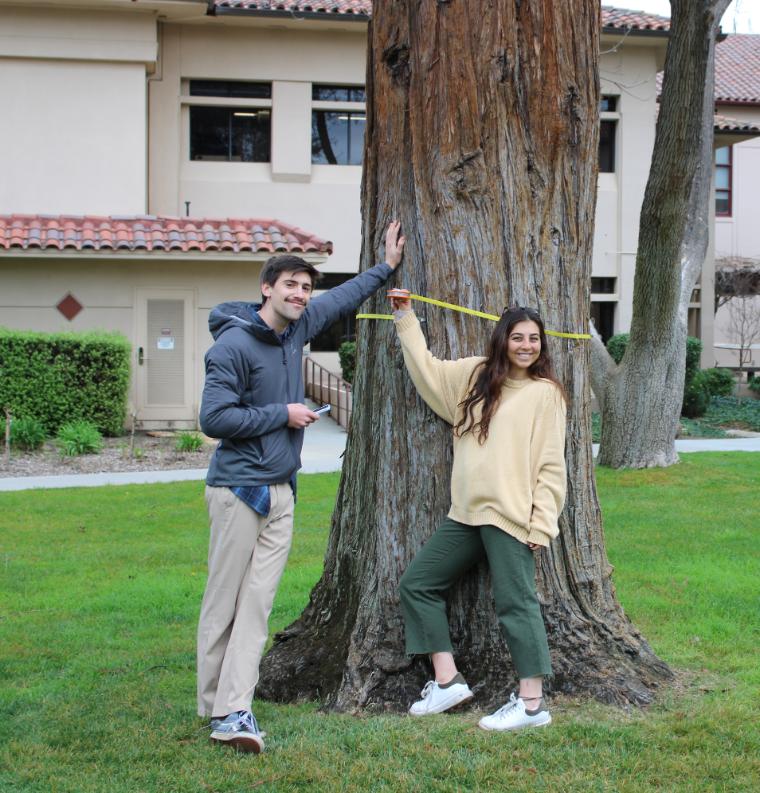 2 Tree Trackers students measuring a by a tree with measuring tape while smiling