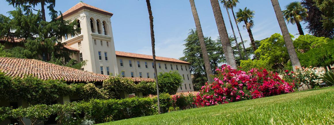 Large building with a white tower, palm trees, and gardens in a sunny setting.