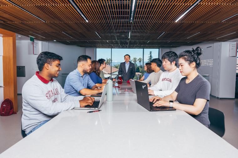 8 students sitting at a long table, four on each side, with a professor standing at the end giving a lecture.