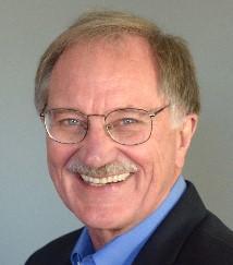 A smiling person wearing glasses, a blue shirt, and a dark jacket.