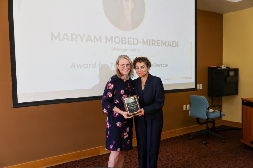 Two women standing side by side holding an award plaque in front of a presentation screen for a Bioengineering teaching excellence award