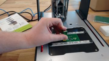 A hand holding a green circuit board near a 3D printer.